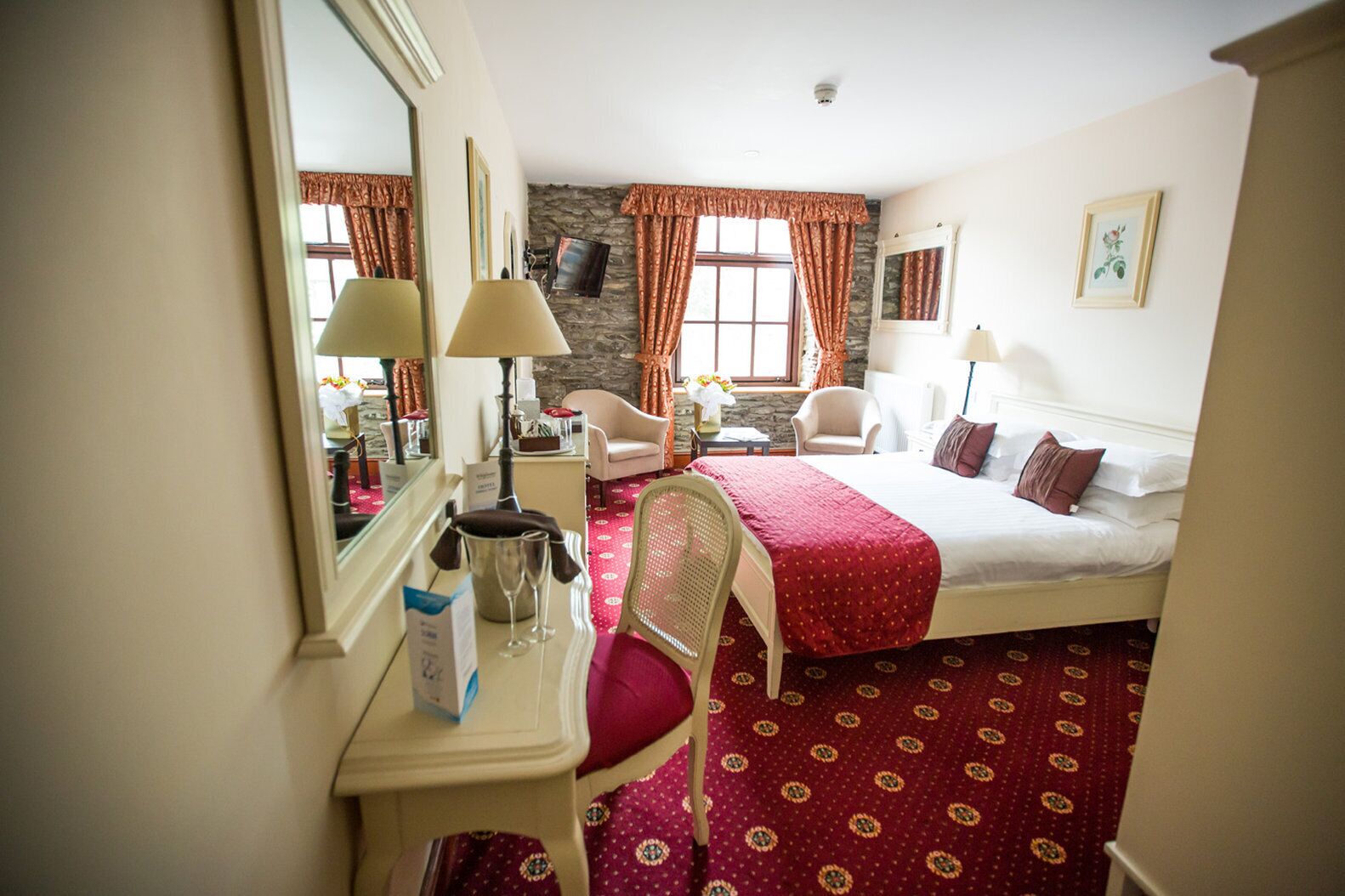 A hotel room with a bed, a desk with a mirror, a chair, a small table with a vase of flowers, and a window with curtains.