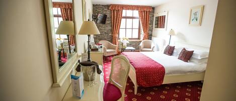 A hotel room with a bed, a desk with a mirror, a chair, a small table with a vase of flowers, and a window with curtains.