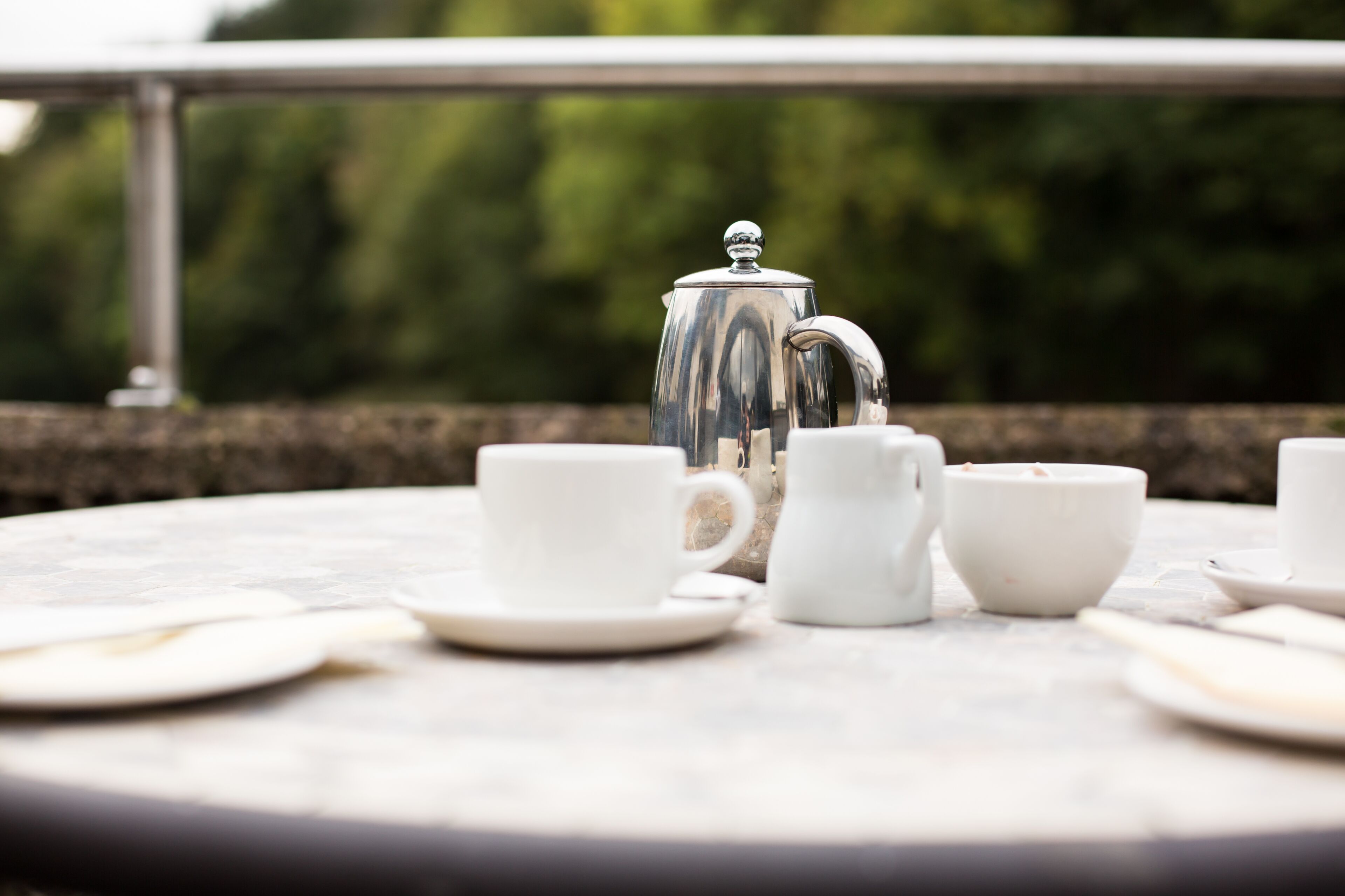 Une table ronde avec une théière en argent et des tasses et soucoupes blanches.