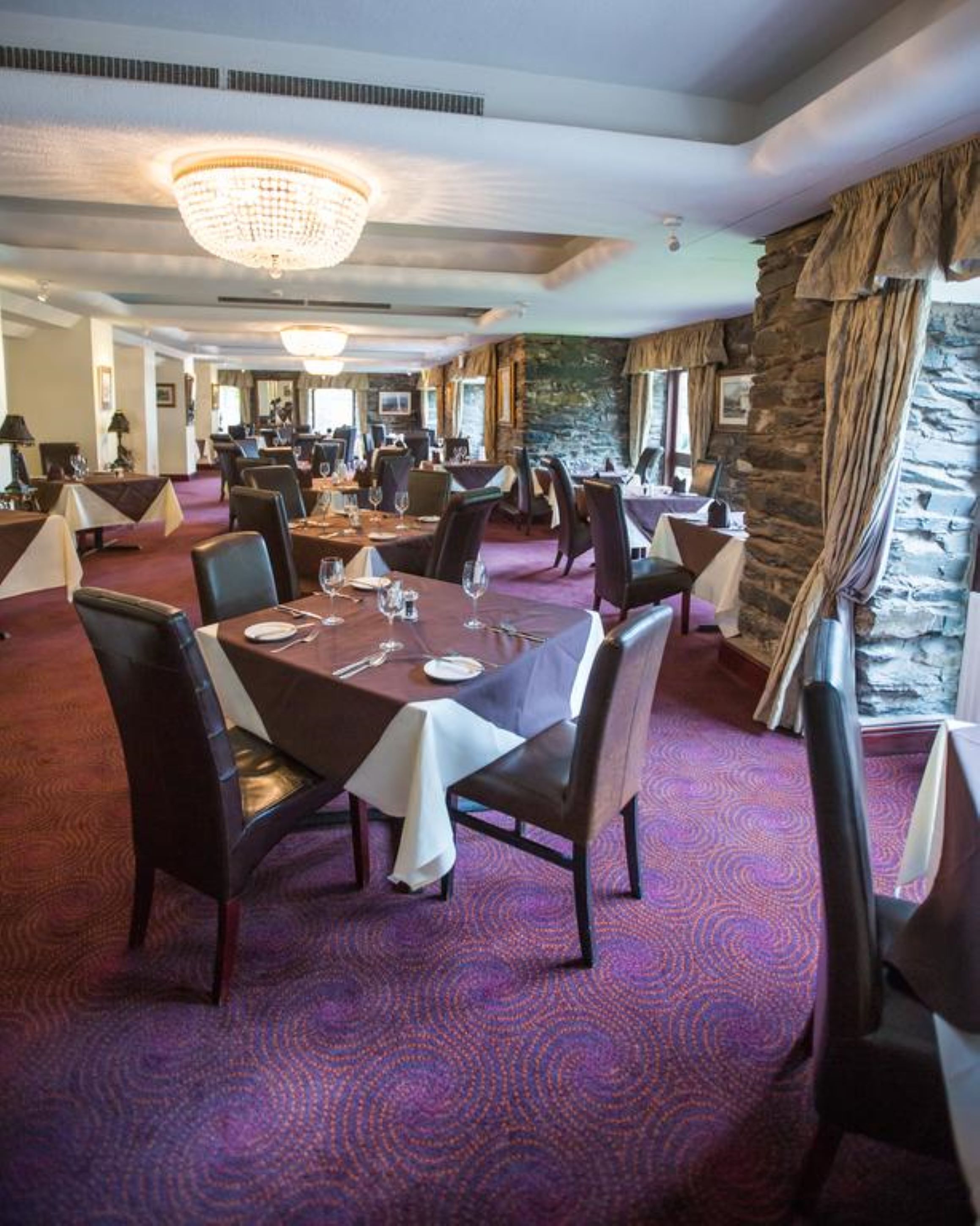 Un restaurante con varias mesas, sillas oscuras y una araña. Las mesas están dispuestas con platos, vasos y cubiertos. La alfombra tiene un diseño circular, y hay una pared de piedra con una ventana en un lateral.