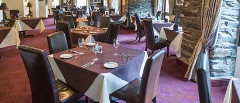 Un restaurante con varias mesas, sillas oscuras y una araña. Las mesas están dispuestas con platos, vasos y cubiertos. La alfombra tiene un diseño circular, y hay una pared de piedra con una ventana en un lateral.