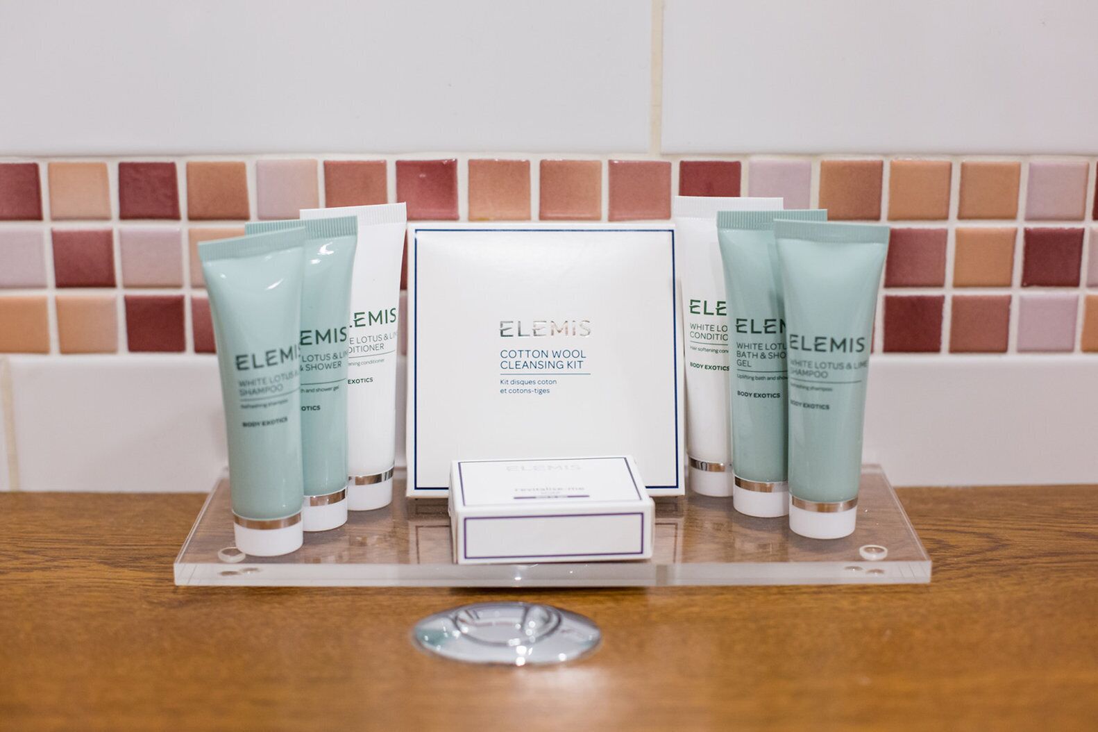 A display of Elemis skincare products including a cleansing kit, body lotion, and shampoo, arranged on a table with a tiled backsplash in the background.