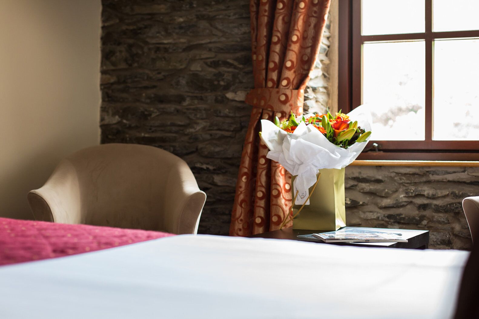 A hotel room with a stone wall, a window with curtains, a bed with a white bedspread, a chair, and a bouquet of flowers.
