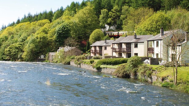 A riverside hotel with multiple buildings, a river flowing in the foreground, and a forested hillside in the background. - Whitewater Spa & Leisure Club (Ulverston)