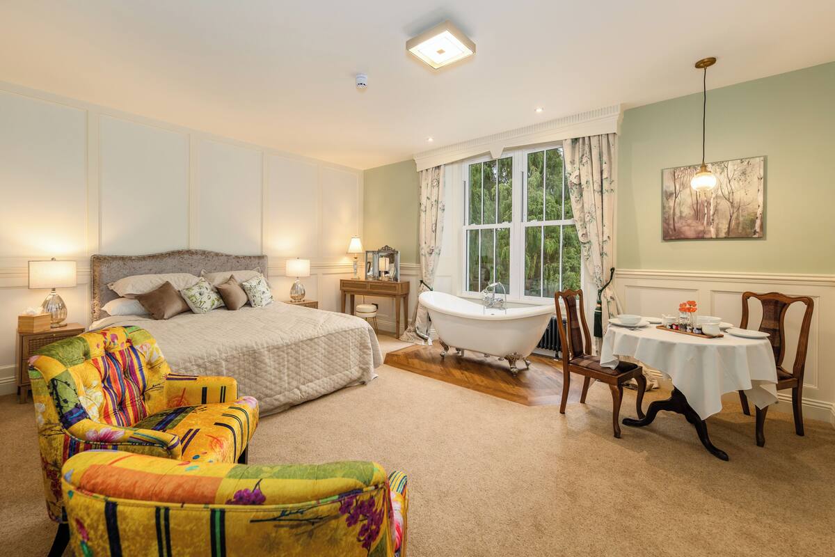 A bedroom with a large bed, a colorful armchair, a bathtub, and a dining table with chairs.