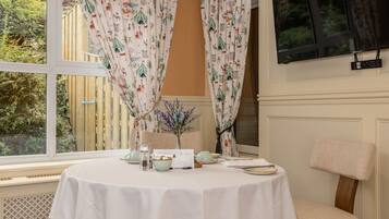 Una habitación con una mesa redonda dispuesta para una comida, una silla, una ventana con cortinas y vistas a follaje exterior.