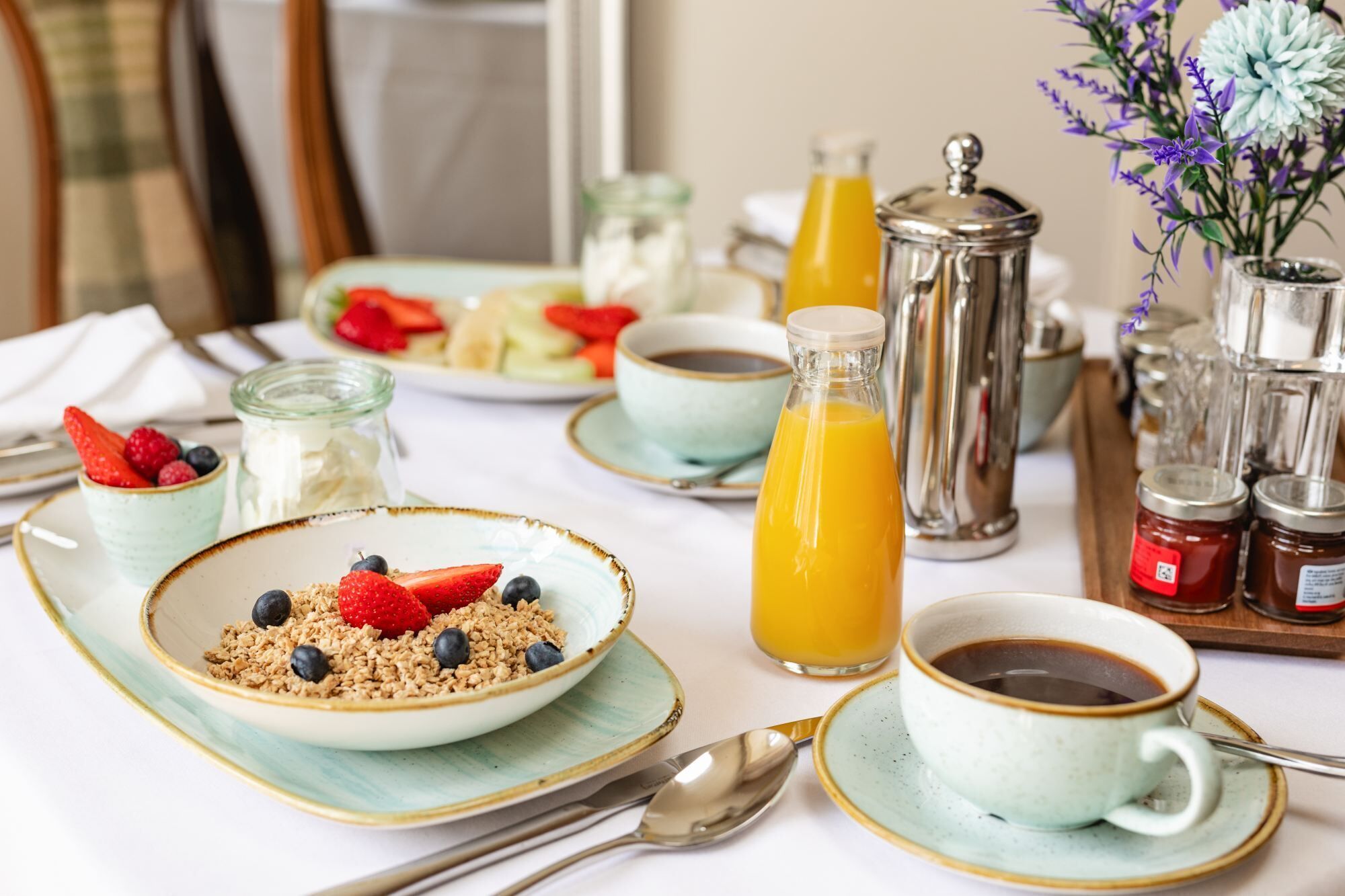 Une table mise pour le petit-déjeuner avec une assiette de granola, des fruits rouges, un verre de jus d’orange, une tasse de café et un pot de confiture.