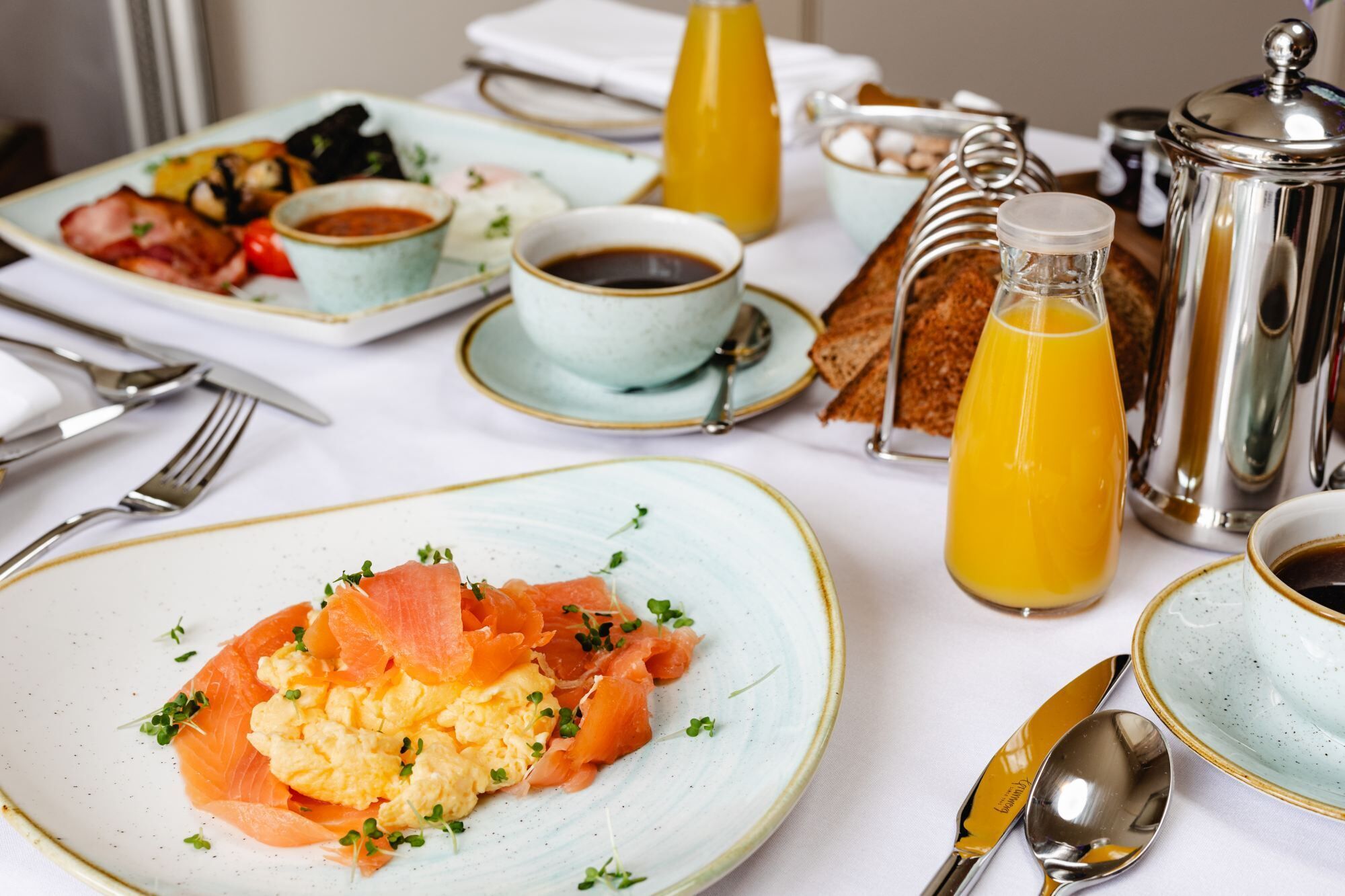 A breakfast setting with plates of smoked salmon and scrambled eggs, a bottle of orange juice, and a coffee pot.
