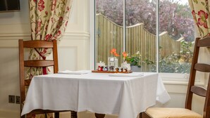 A dining area with a table set for two, a wooden chair, and a window with a view of a garden. - Grange Manor (Grange-over-Sands)