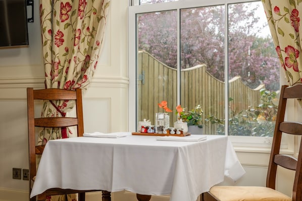 A dining area with a table set for two, a wooden chair, and a window with a view of a garden. - Grange Manor (Grange-over-Sands)