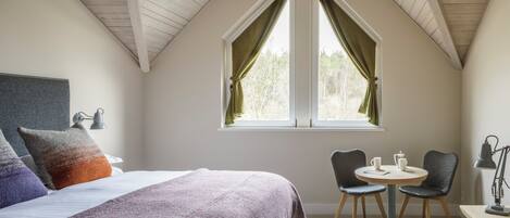 A bedroom with a large bed, a window with curtains, a desk with chairs, and a lamp.