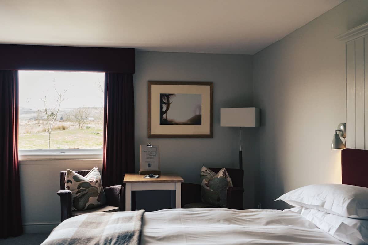 Habitación de hotel con cama, escritorio, silla, lámpara y ventana con cortinas.