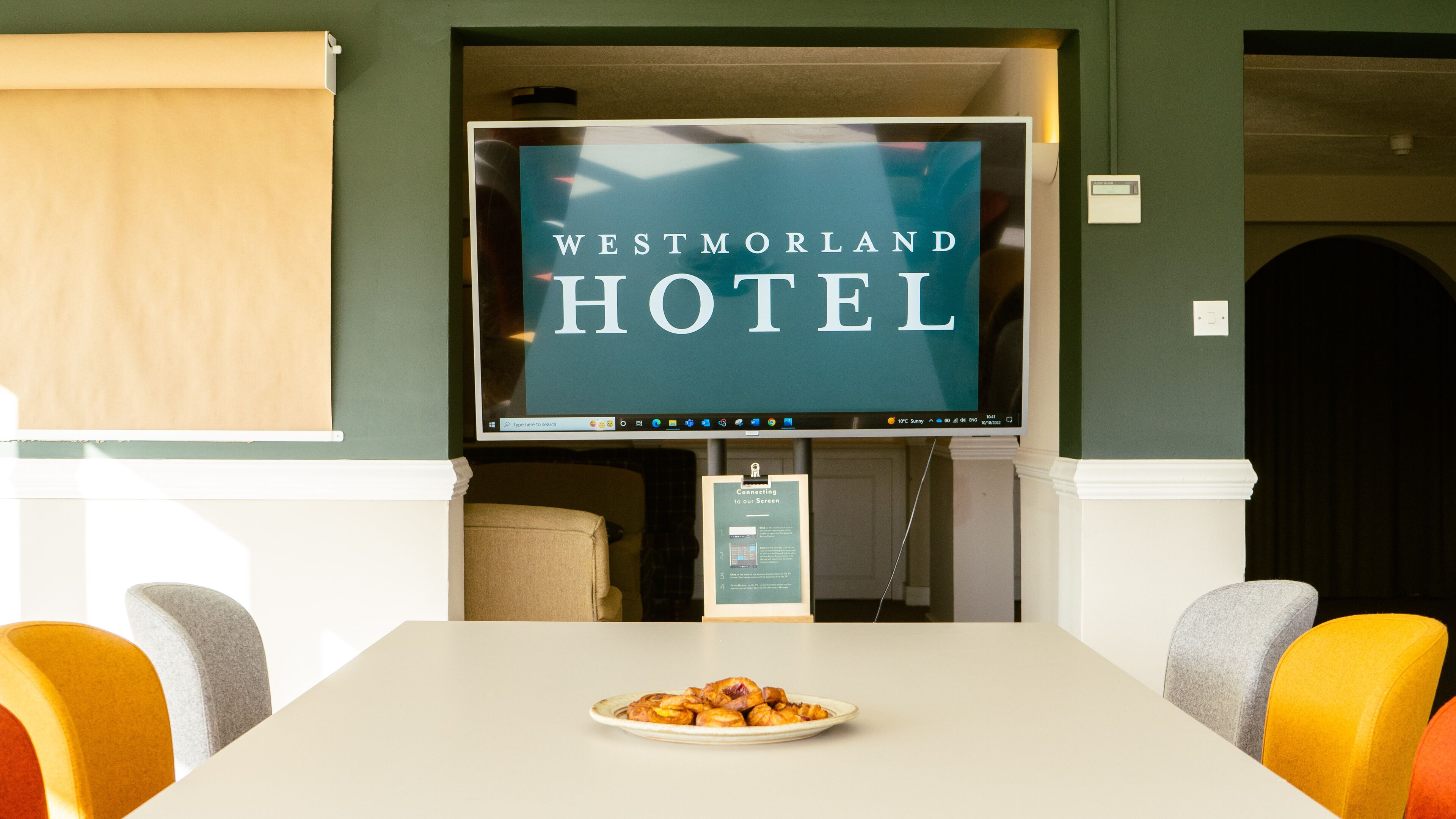 Recepção do Westmorland Hotel, com uma tela grande exibindo o nome do hotel, uma mesa com uma bandeja de doces e cadeiras coloridas.