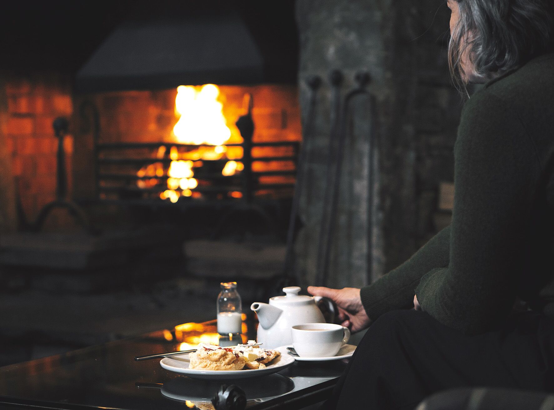 A person sitting at a table with a plate of food, a cup, and a teapot, in front of a fireplace.