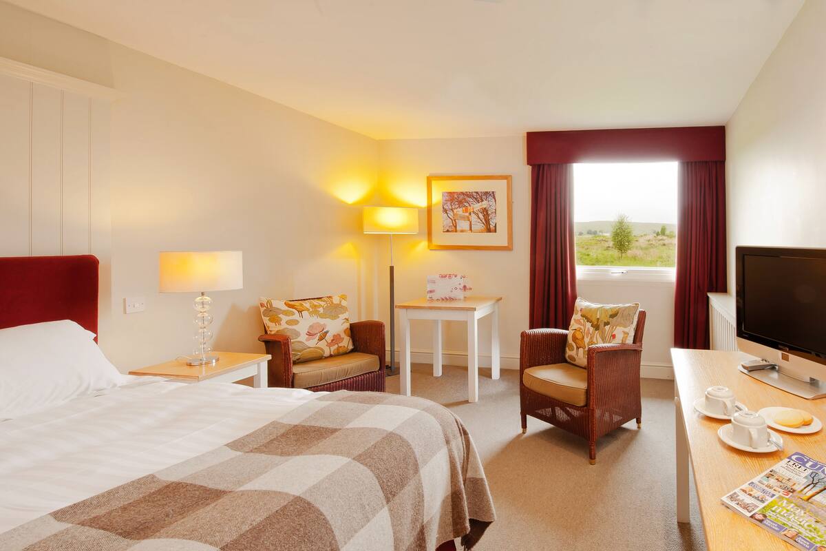 Habitación de hotel con cama, escritorio con computadora, silla, mesita y ventana con cortinas.