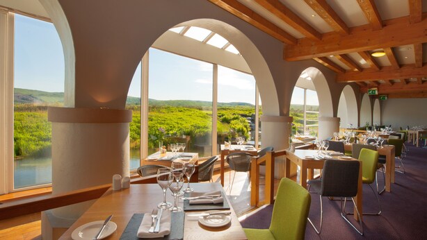 Restaurante con ventanales altos y arqueados que ofrece vistas a colinas verdes. Las mesas están dispuestas con copas de vino y platos.
