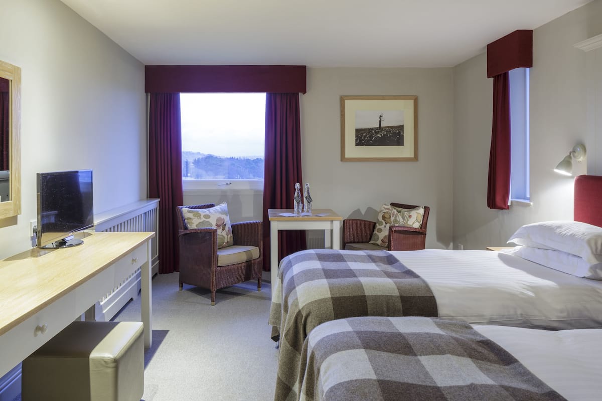 Habitación de hotel con cama, escritorio con computadora, silla, mesita y ventana con cortinas.