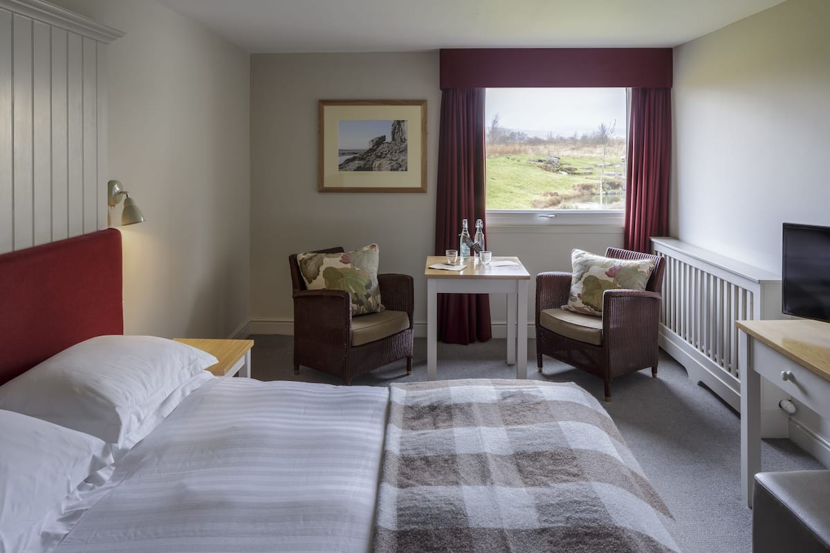 Habitación de hotel con cama, dos sillones, una mesita de noche con lámpara, televisor y ventana con vistas al campo.