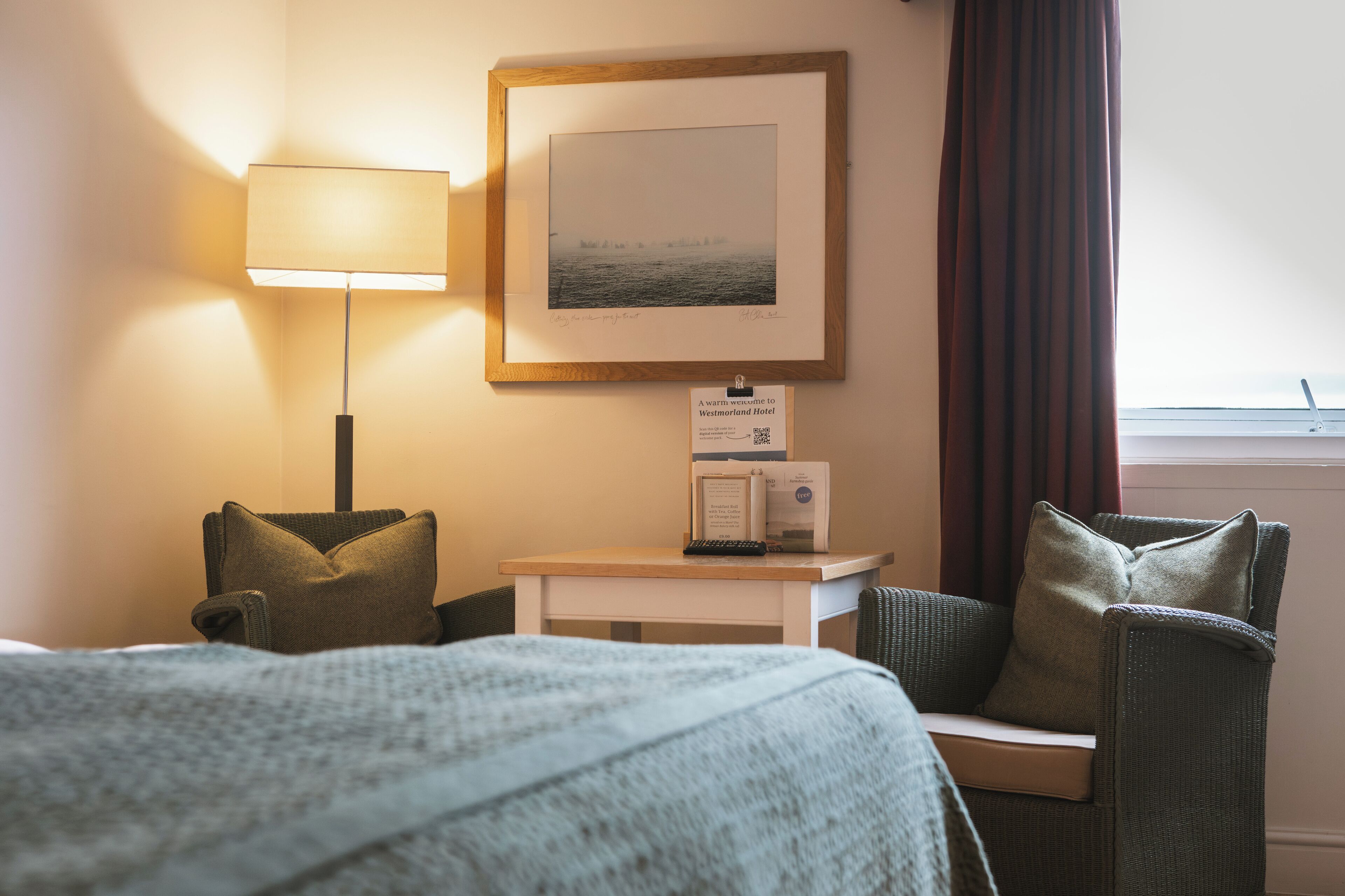 A hotel room with a bed, a desk with a lamp, a framed picture on the wall, and a window with curtains.