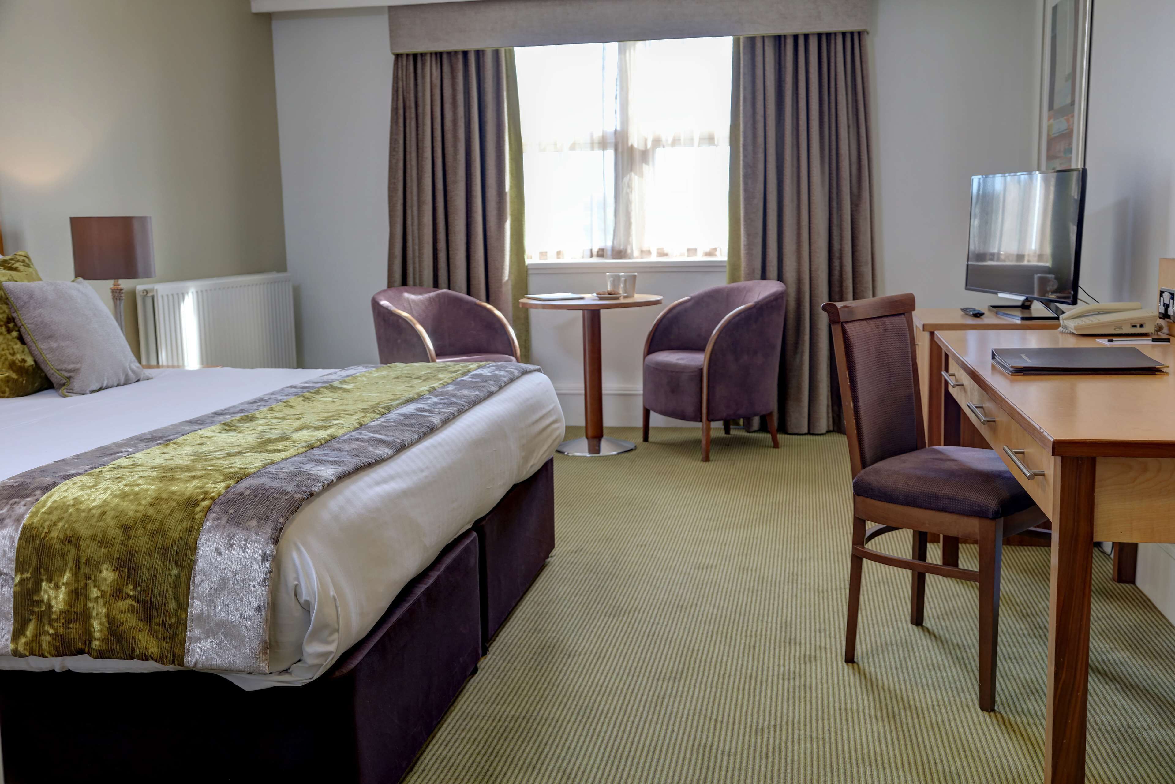 A hotel room with a bed, a desk, two chairs, a television, and a window with curtains.