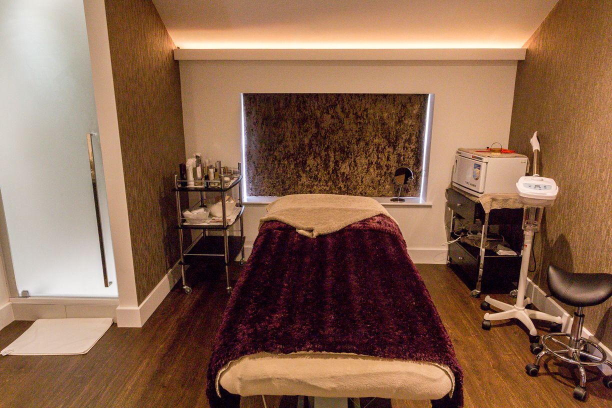 A treatment room with a bed, a window, a shelf with bottles, and a chair.