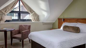A hotel room with a large bed, a chair, a small table, and a window with curtains.