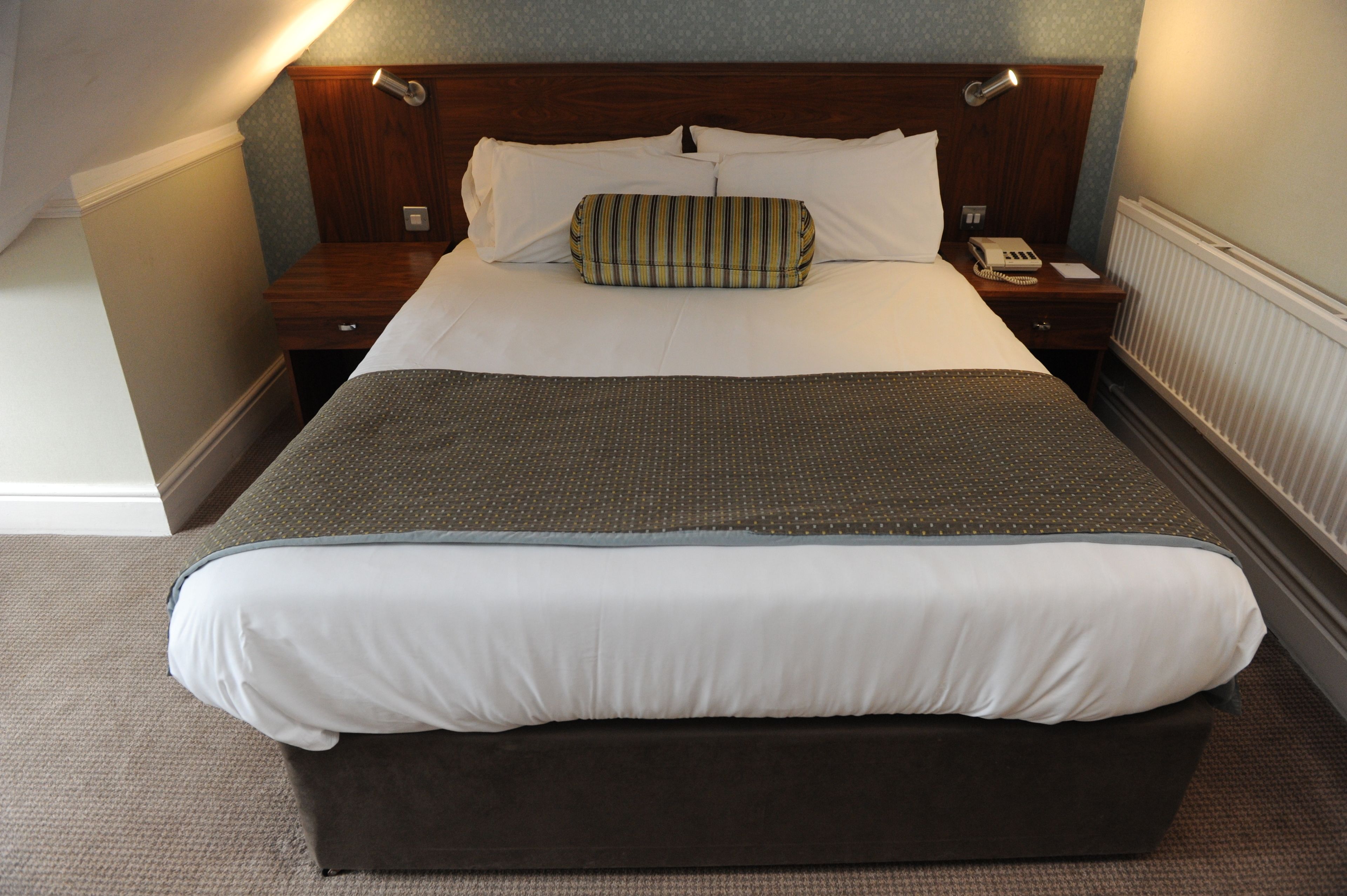 A hotel room with a large bed, a wooden headboard, a bedside table with a lamp, and a telephone.