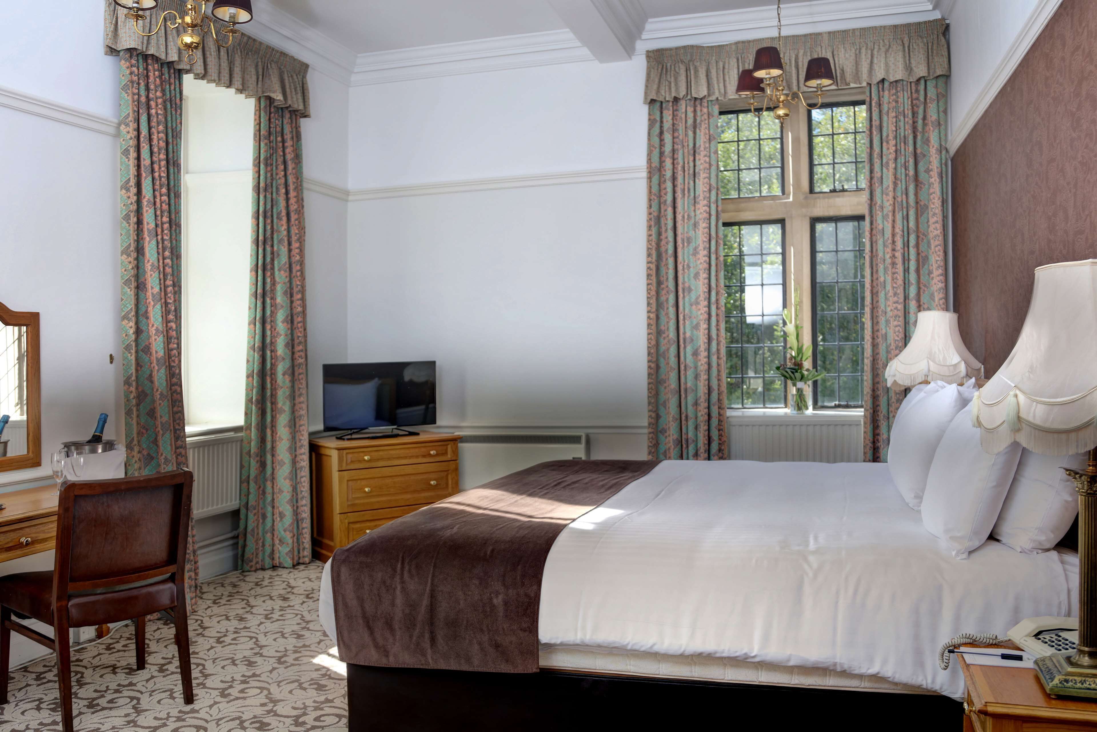 A hotel room with a large bed, a wooden desk, a television, and a window with curtains.