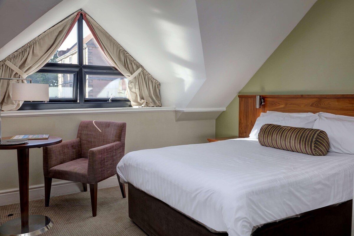 A hotel room with a bed, a chair, a small table, and a window with curtains.