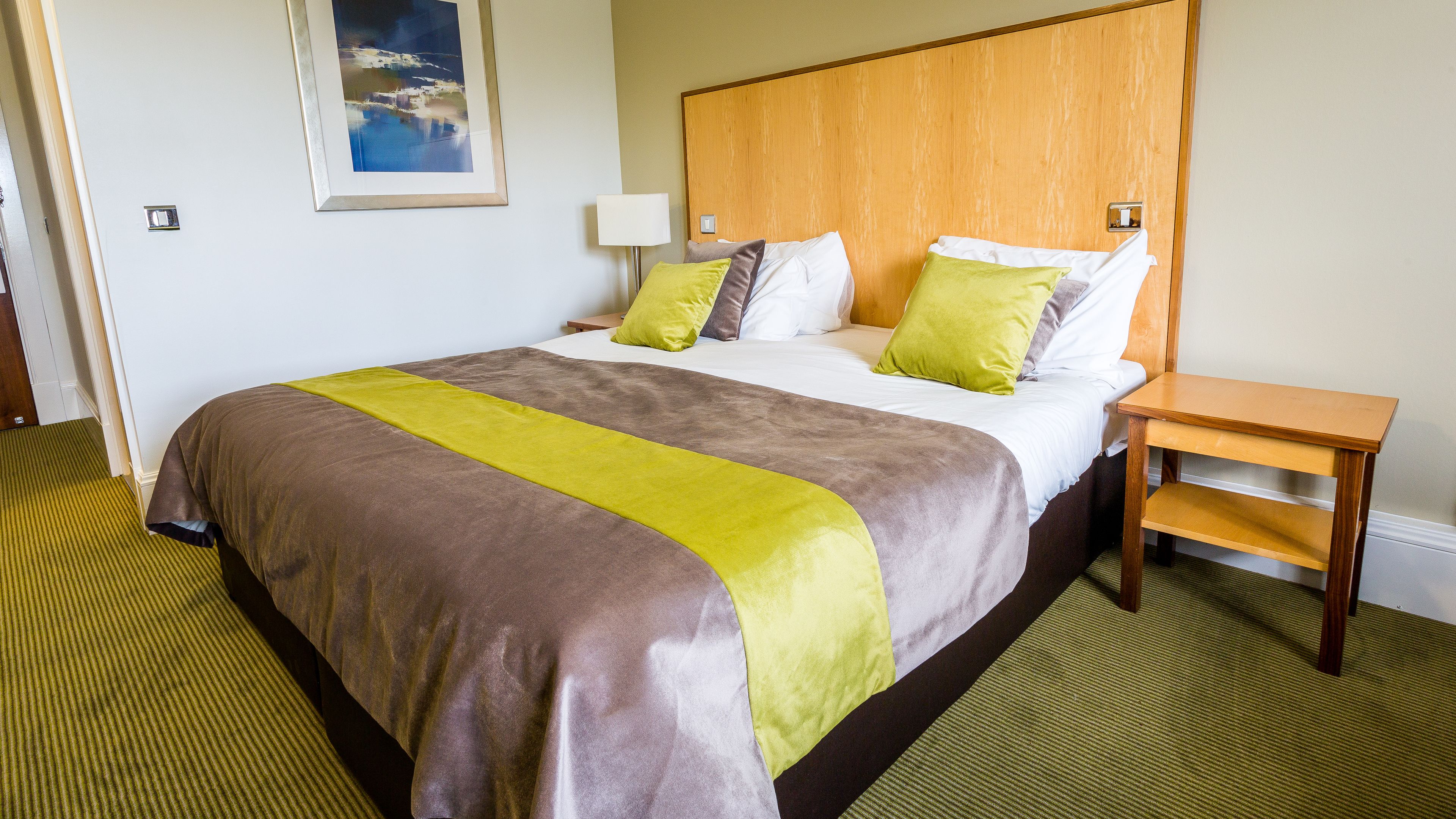 A hotel room with a large bed, a wooden headboard, a nightstand, and a framed picture on the wall.