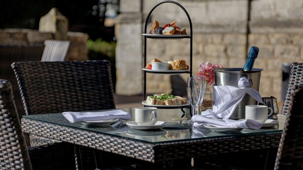 A three-tiered dessert stand with various pastries, a champagne bottle, and a glass of champagne on a table with wicker chairs. - Hellaby Hall Hotel, BW Signature Collection (Rotherham)