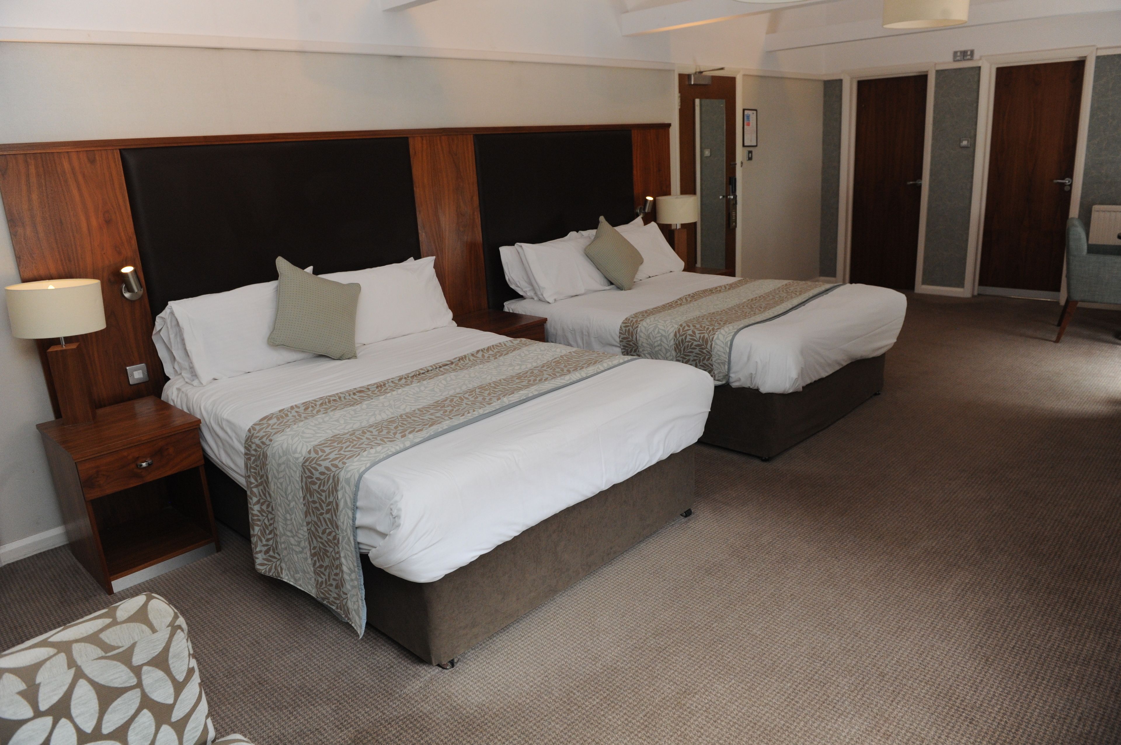 Habitación de hotel con dos camas, cabeceros de madera, mesitas de noche con lámparas y suelo alfombrado.