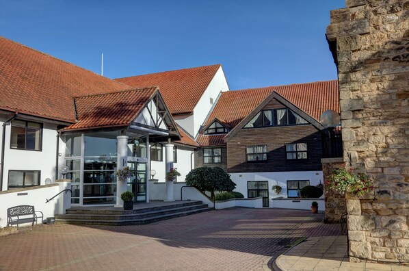 A traditional European-style building with a red-tiled roof, a central entrance with steps, and a stone wall on the right.