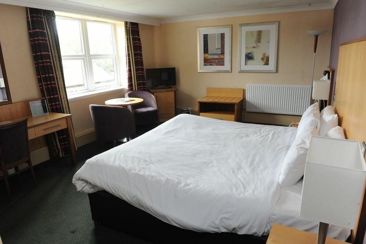 A hotel room with a large bed, a desk with a chair, a small table, a TV, and two framed pictures on the wall.