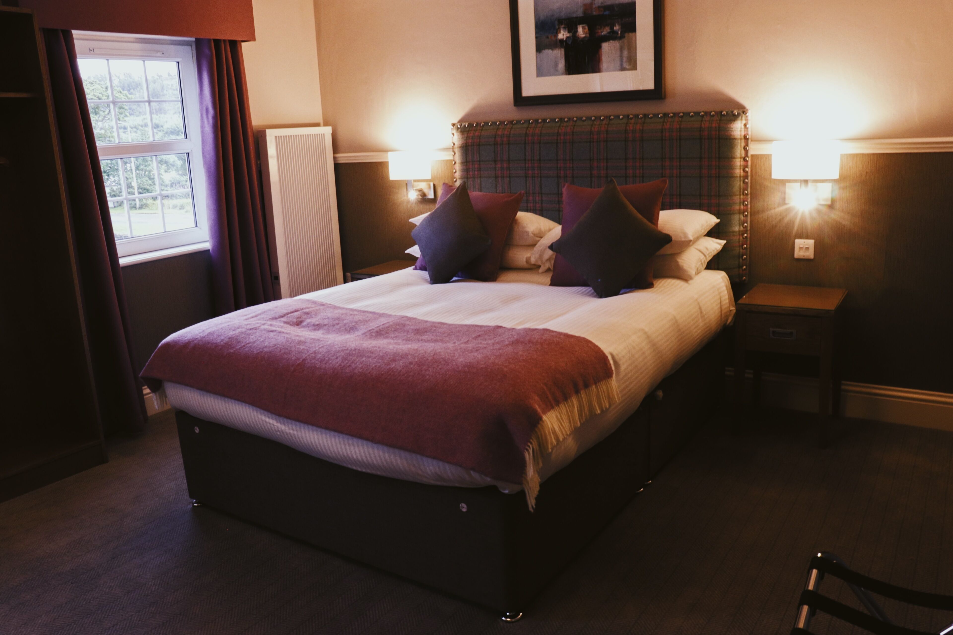 A neatly made bed with a plaid headboard, two bedside tables with lamps, and a window with curtains.