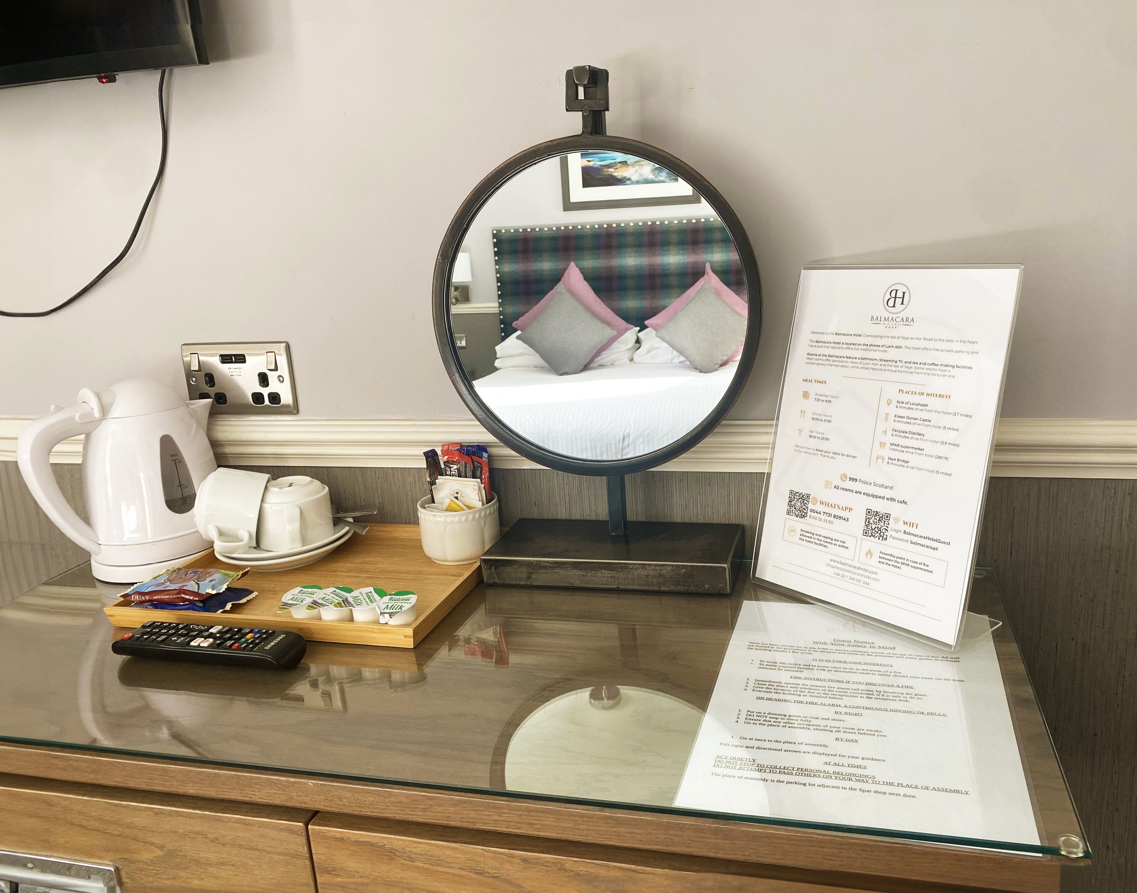 A hotel room with a bed, a mirror, a kettle, a cup, a tray with snacks, a remote control, and a welcome brochure.