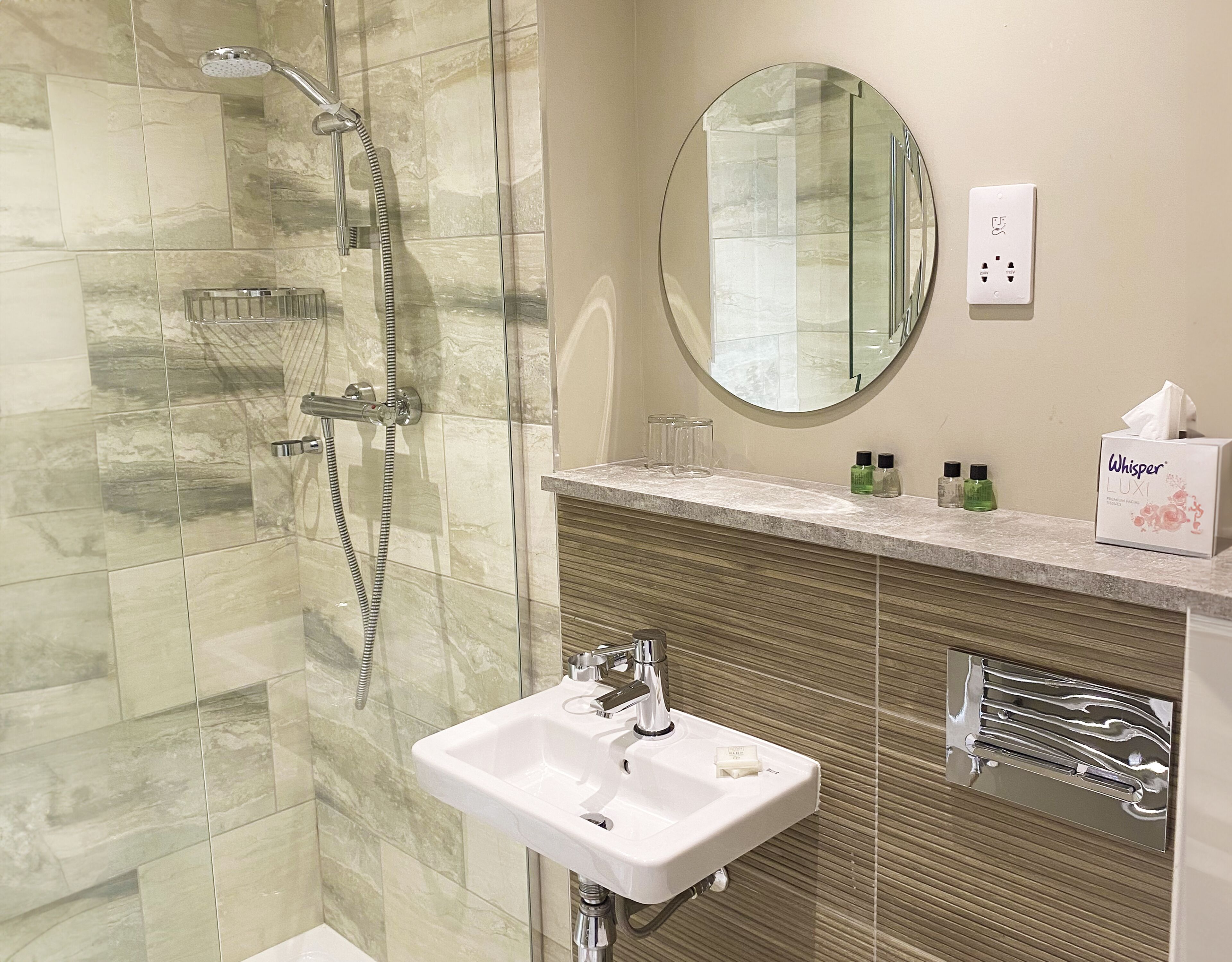 A modern bathroom with a glass-enclosed shower, a white sink, a vanity with toiletries, and a round mirror.