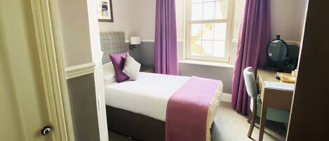 A hotel room with a bed, a desk, a chair, a window with curtains, a lamp, and a framed picture on the wall.