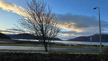 Une vue prise au bord de la route, avec un arbre, un mur de pierres, un panneau indiquant un parking et une montagne en arrière-plan.