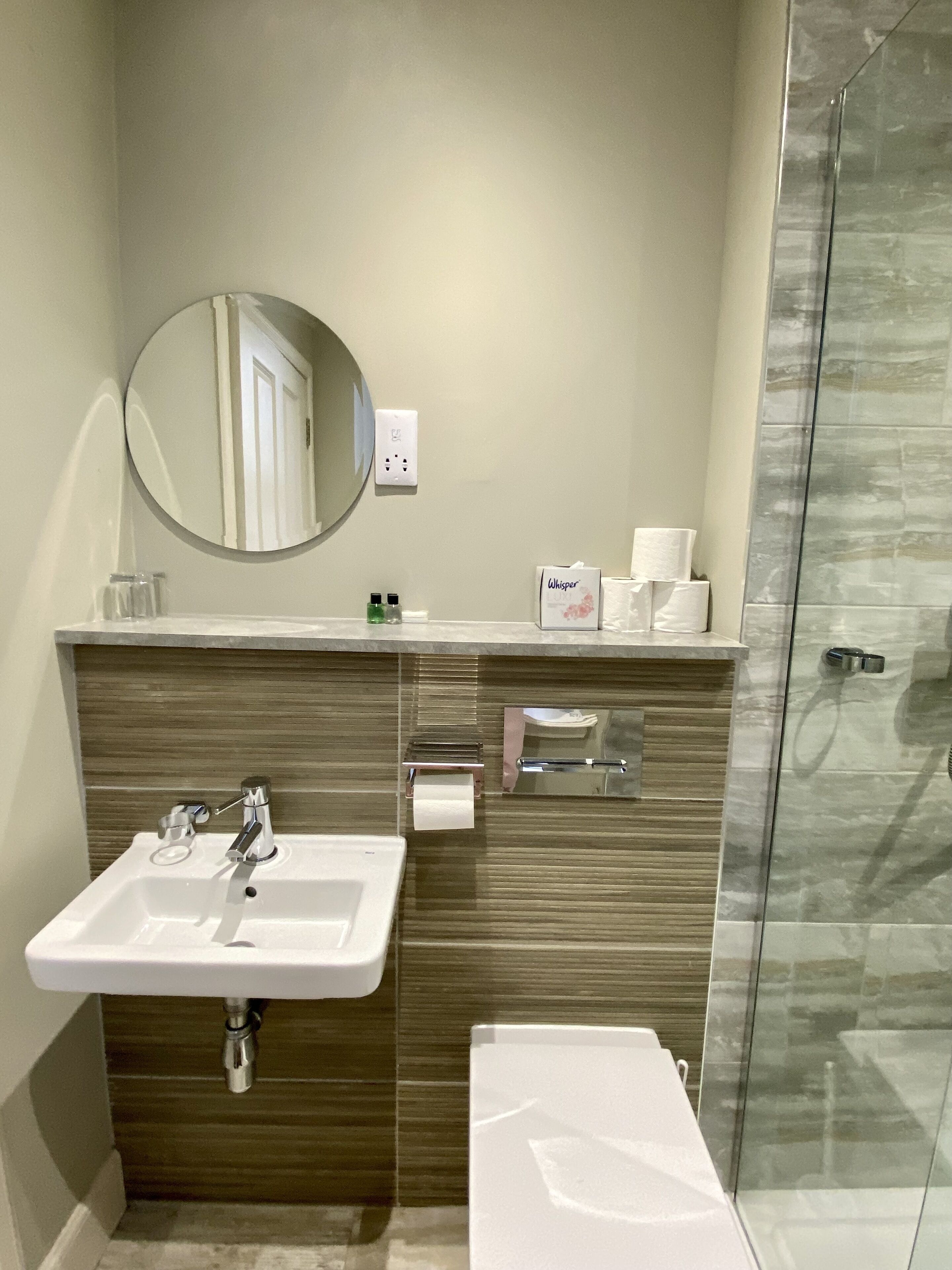 A modern bathroom with a white sink, a mirror, a towel dispenser, and a glass shower enclosure.