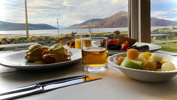A table with plates of food, a glass of beer, and a bowl of fruit, with a scenic view of mountains and a lake through a window. - Balmacara Hotel (Kyle)