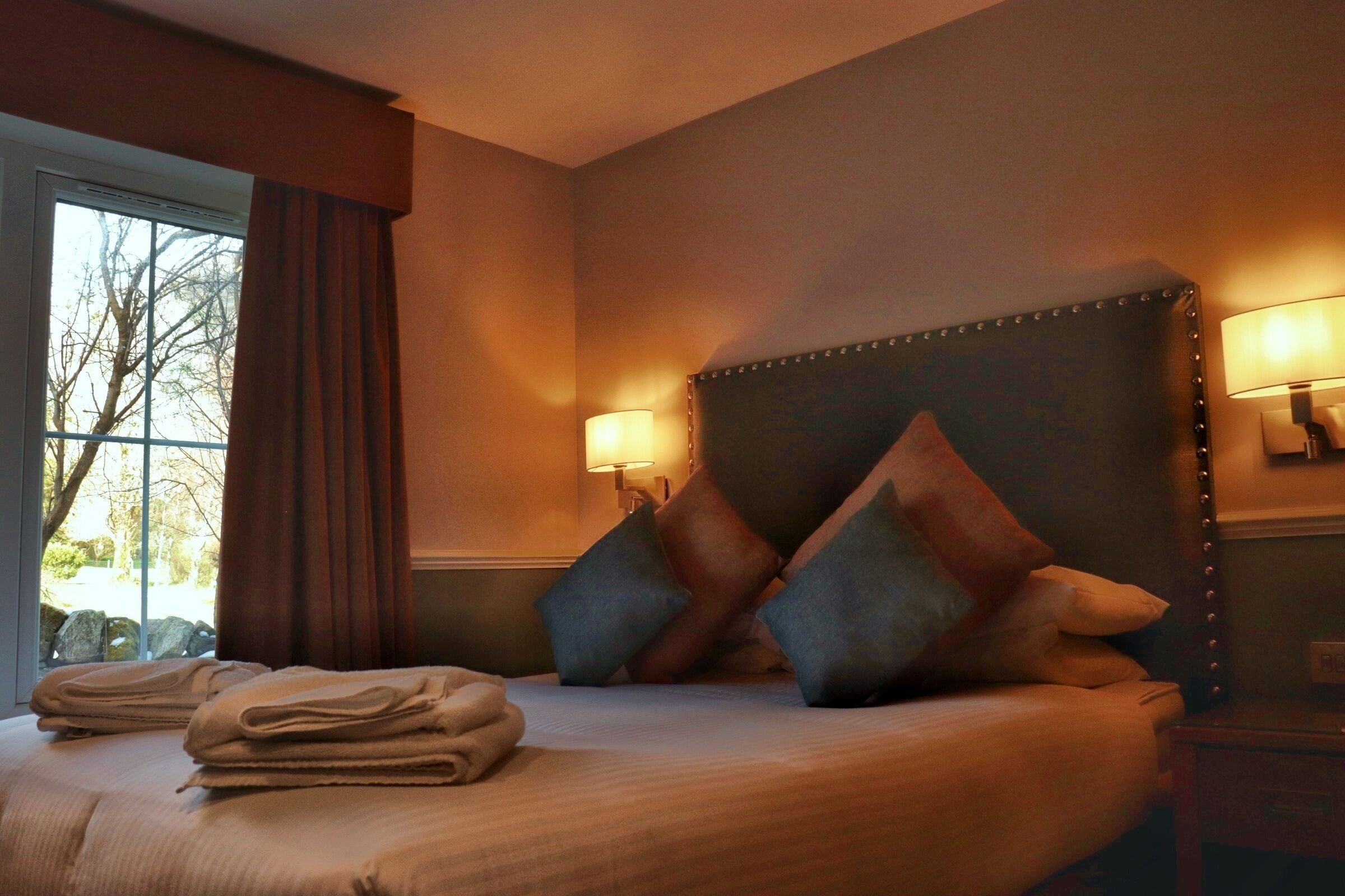 Une chambre d’hôtel avec un grand lit, deux lampes de chevet, une fenêtre avec des rideaux et une vue sur les arbres à l’extérieur.