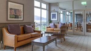 A hotel lobby with a sofa, armchair, and table, large windows, and a view of the outdoors. - Balmacara Hotel (Kyle)