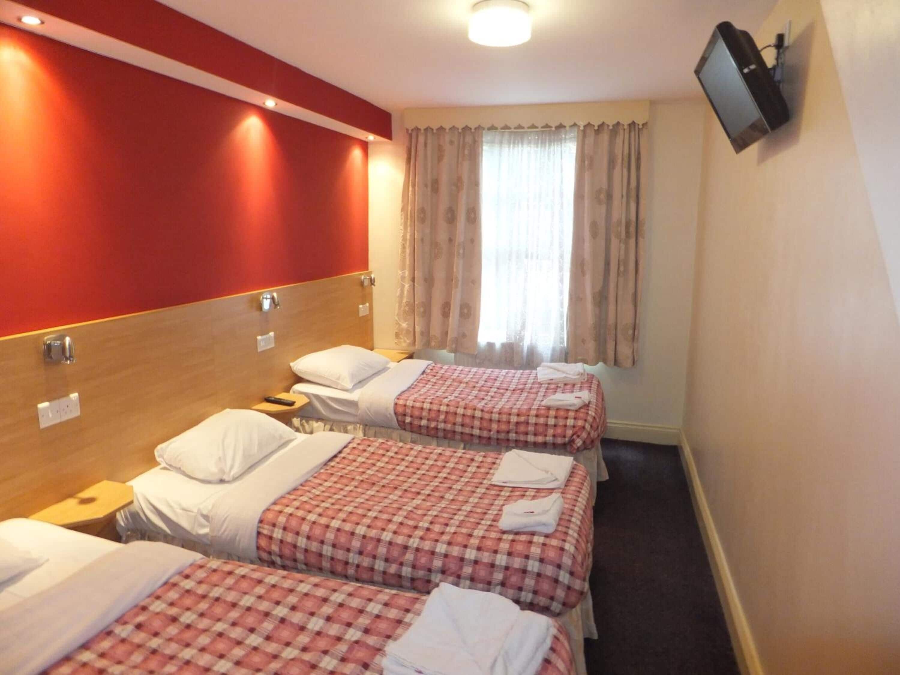 A hotel room with three single beds, each with a red and white checkered bedspread and white pillows. There is a television mounted on the wall to the right, and a window with beige curtains.