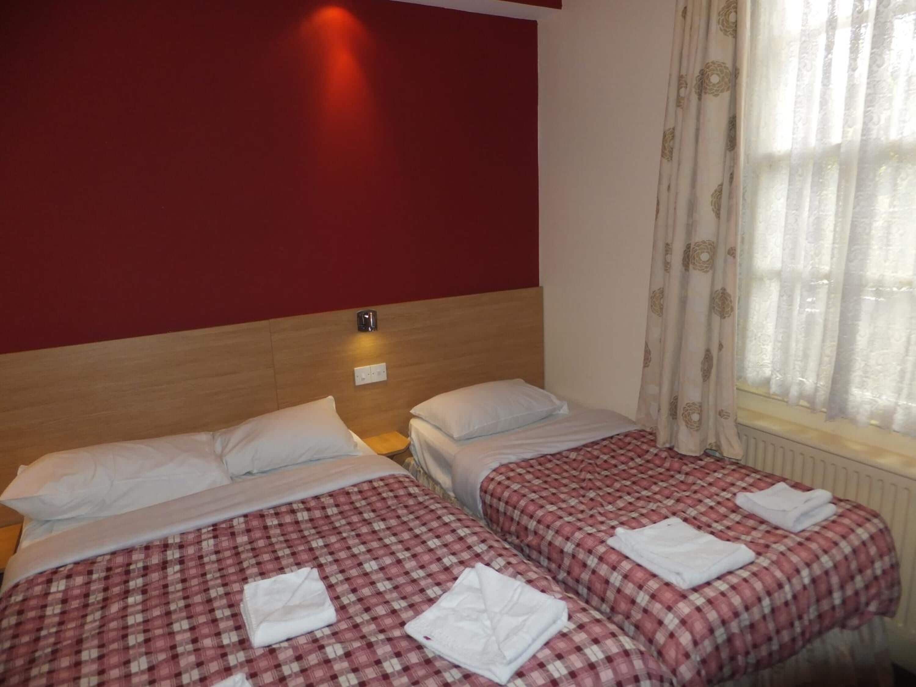 Habitación de hotel con dos camas, cada una con colcha a cuadros rojos y blancos y almohadas blancas. Entre las camas, un mesita de noche con lámpara, y una ventana con cortinas traslúcidas.