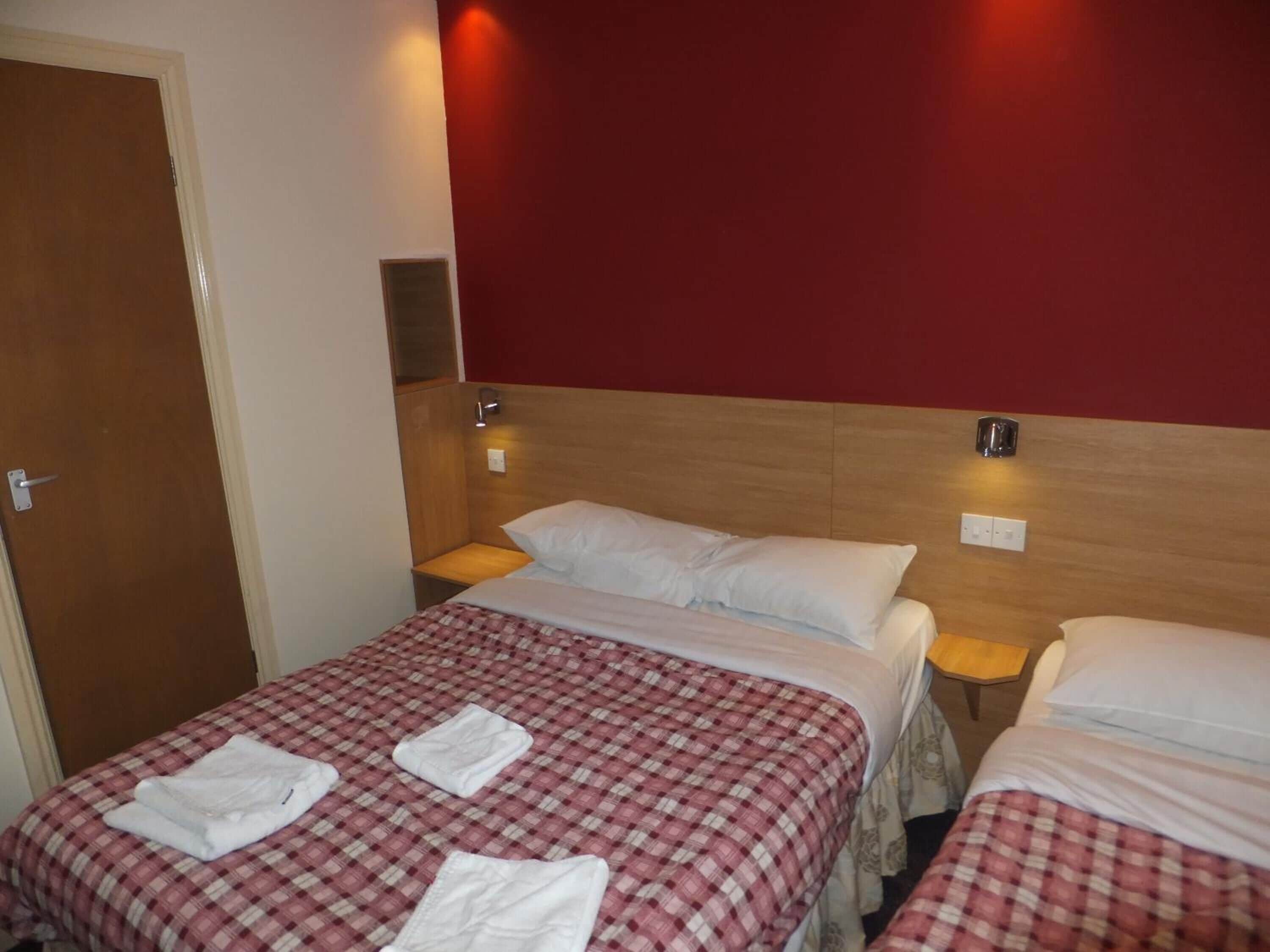 Ein Hotelzimmer mit zwei Betten, jedes mit einer rot-weißen Karodecke und weißen Kissen. Es gibt zwei Nachttische mit Lampen, ein Holz-Kopfteil und eine Tür.