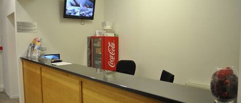 Uma área de recepção de hotel com um balcão de madeira, uma televisão fixada na parede e um refrigerador de Coca-Cola.