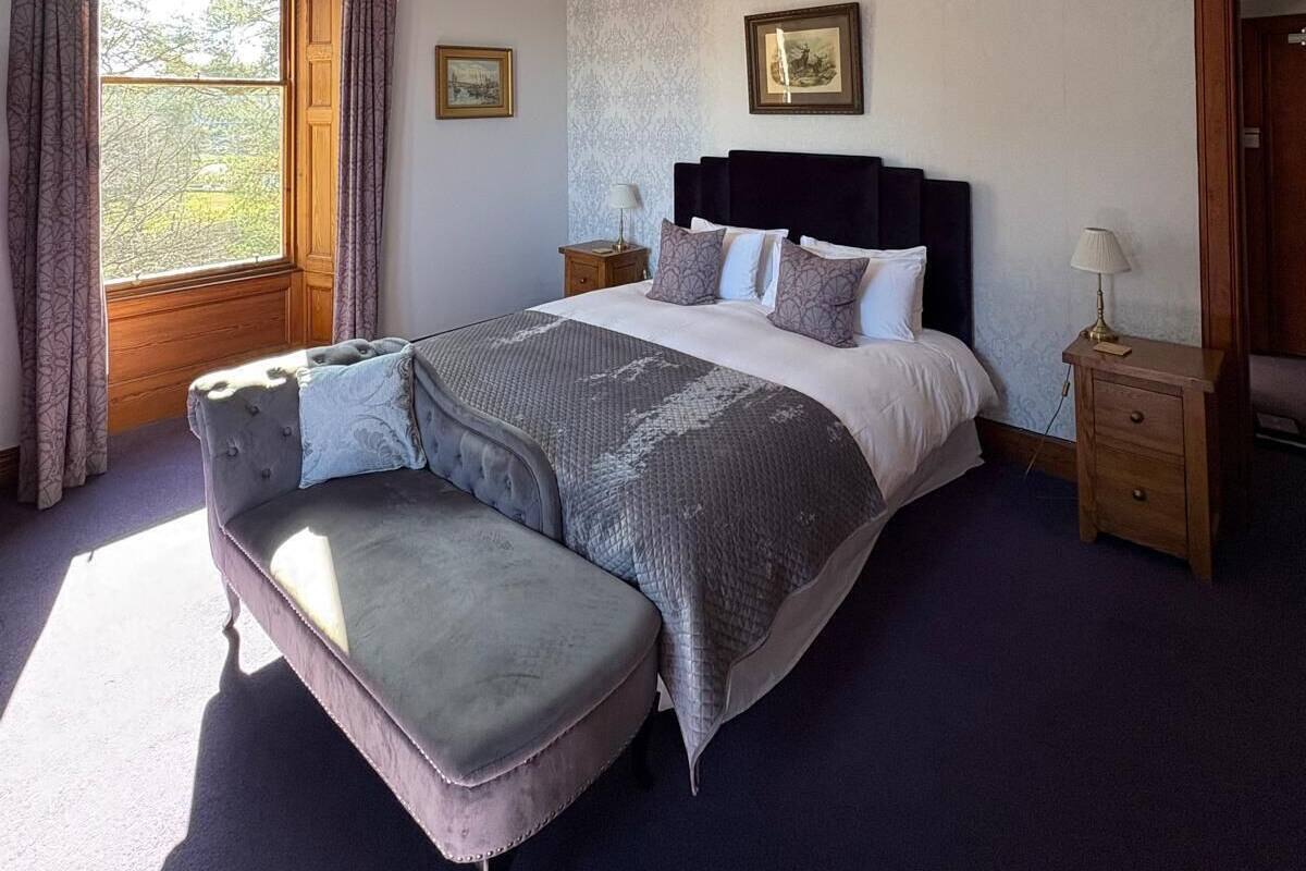 Een slaapkamer met een groot bed, een bankje, twee nachtkastjes, een raam met gordijnen en een plafondlamp.