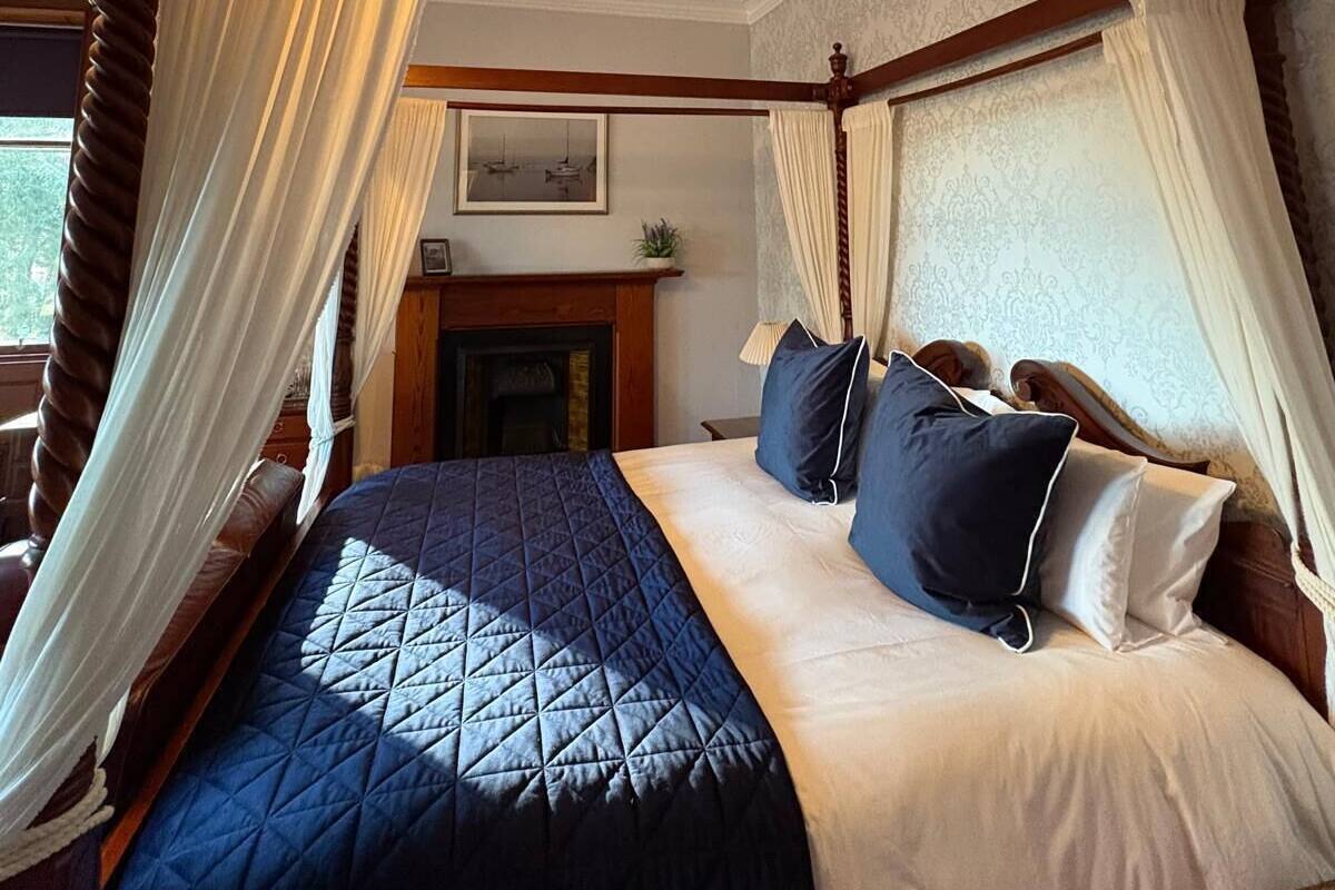 Een hemelbed met een gewatteerde sprei, witte en blauwe kussens en een baldakijn met witte gordijnen.