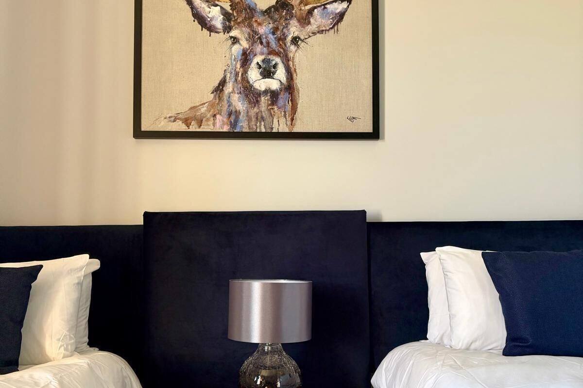 Een hotelkamer met twee bedden, een houten nachtkastje met een lamp, en een lijst met daarop een hertenbeeld.